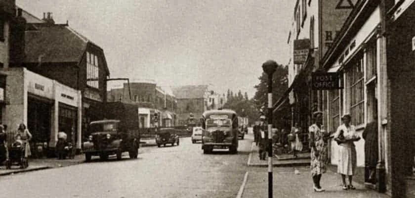 A black-and-white photo of a mid-20th-century Swanley street scene, featuring vintage cars, a bus, pedestrians, and shopfronts like the post office. Two women stand on the pavement in the foreground, capturing Swanley heritage.