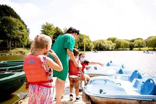 A woman and three children wearing life jackets stand on a jetty by pedalos at Swanley Park on a sunny day, preparing to board. Trees and water are visible in the background.