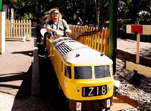Wearing sunglasses, a person rides a small, yellow miniature train on the outdoor tracks of New Barn Railway in Swanley, with trees and a wooden fence in the background.