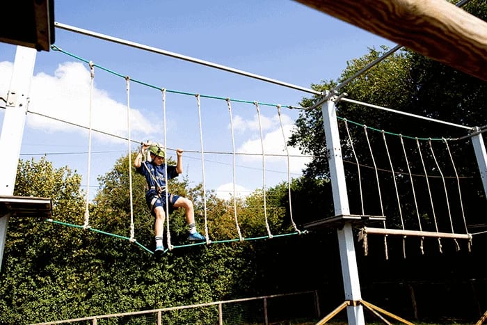 A child wearing a helmet and harness navigates a high ropes course outdoors at Swanley Park, holding onto ropes for balance, with trees and a blue sky in the background—a fantastic destination for family attractions and outdoor activities.