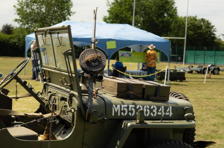 A vintage military jeep with the number M556344 is parked on grass near a blue canopy tent. People are gathered under the tent at an outdoor event on a sunny day.